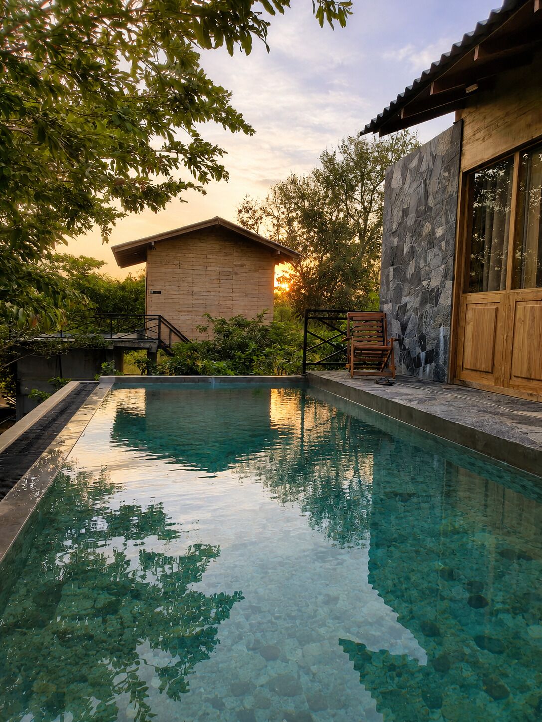 A swimming pool with clear water, surrounded by greenery and a wooden building with stone walls.