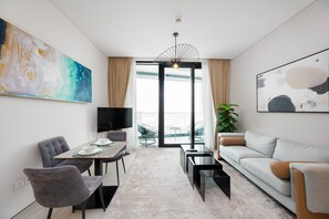 A modern living room with a dining area, a sofa, a coffee table, and a TV. There is a large window with curtains, a plant, and artwork on the walls. - Chic Address JBR Haven w Marina View (Dubai)