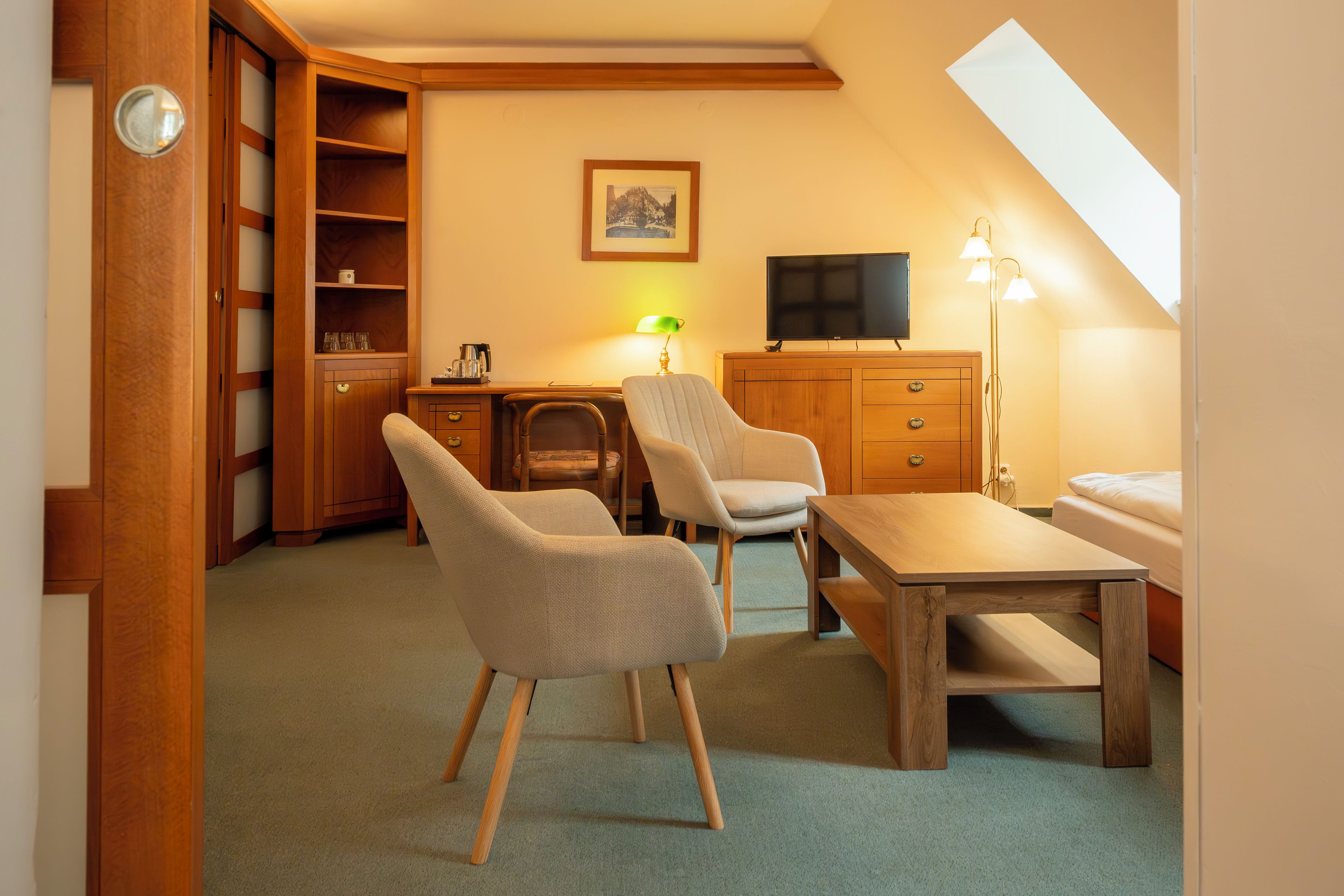 A compact room with a dining table, two chairs, a television, a desk with a lamp, a bookshelf, and a bed.