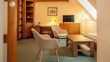 A compact room with a dining table, two chairs, a television, a desk with a lamp, a bookshelf, and a bed.