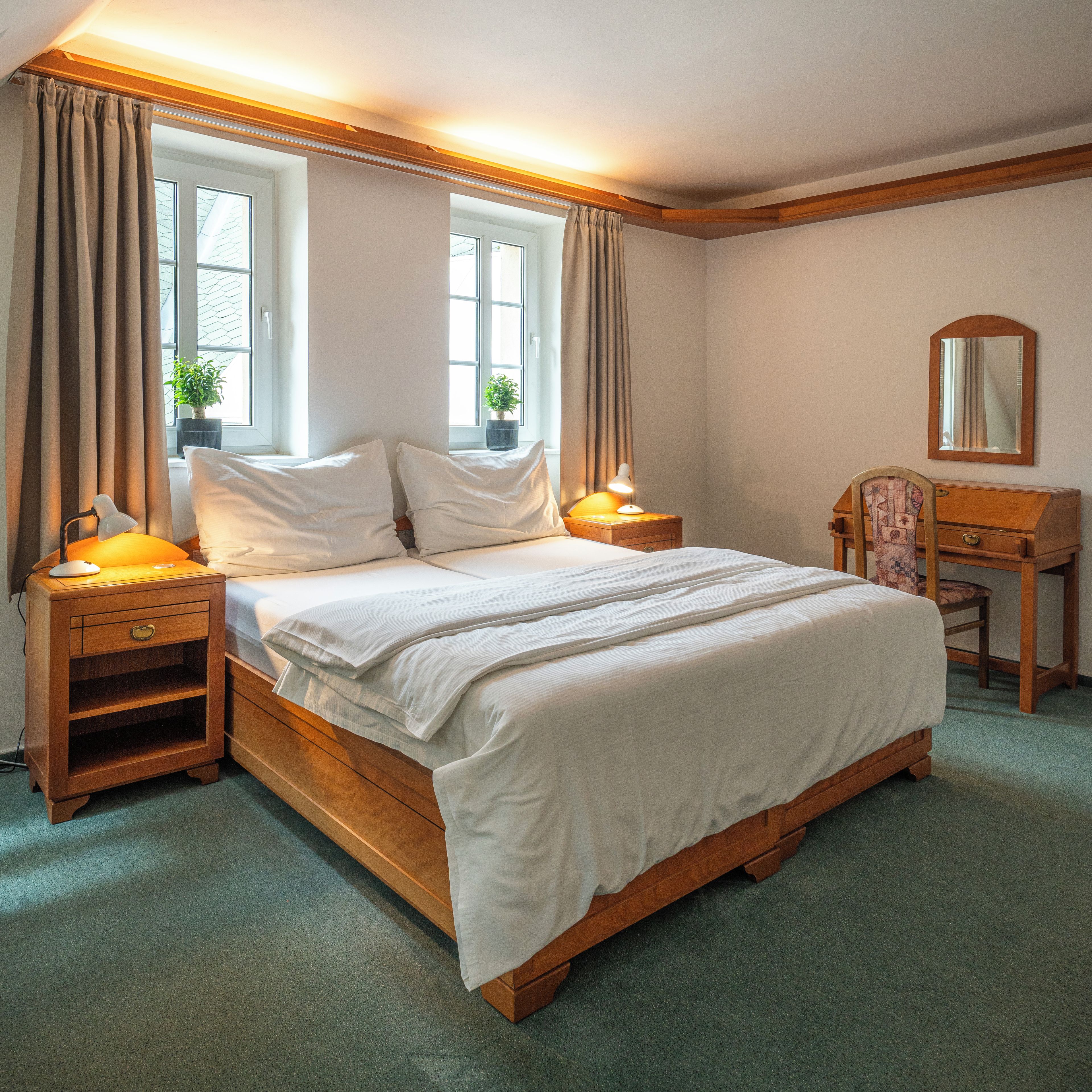 A bedroom with a wooden bed, bedside tables, a nightstand, a dresser, a mirror, and two windows with curtains.