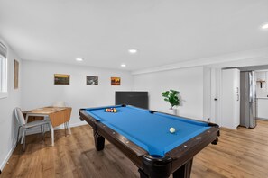 Ein Billardtisch mit Kugeln, ein Fernseher und eine Pflanze in einem Raum mit Holzboden und weißen Wänden.