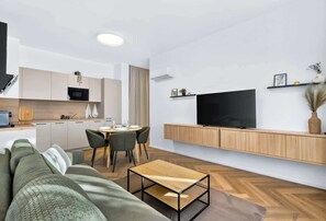 A modern living room with a sofa, a coffee table, a dining area with chairs, a kitchen with cabinets, and a TV mounted on the wall. - Large Family Apartment for 6 - AC & Garage (Bratislava III)