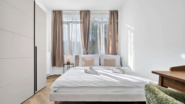 Ein modernes Schlafzimmer mit einem großen Bett, einem Nachttisch mit Lampe und Blick ins Freie durch transparente Vorhänge.