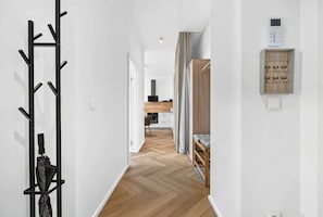 A hallway with a coat rack, a wooden floor, a kitchen area in the distance, and a wall-mounted intercom. - Large Family Apartment for 6 - AC & Garage (Bratislava III)