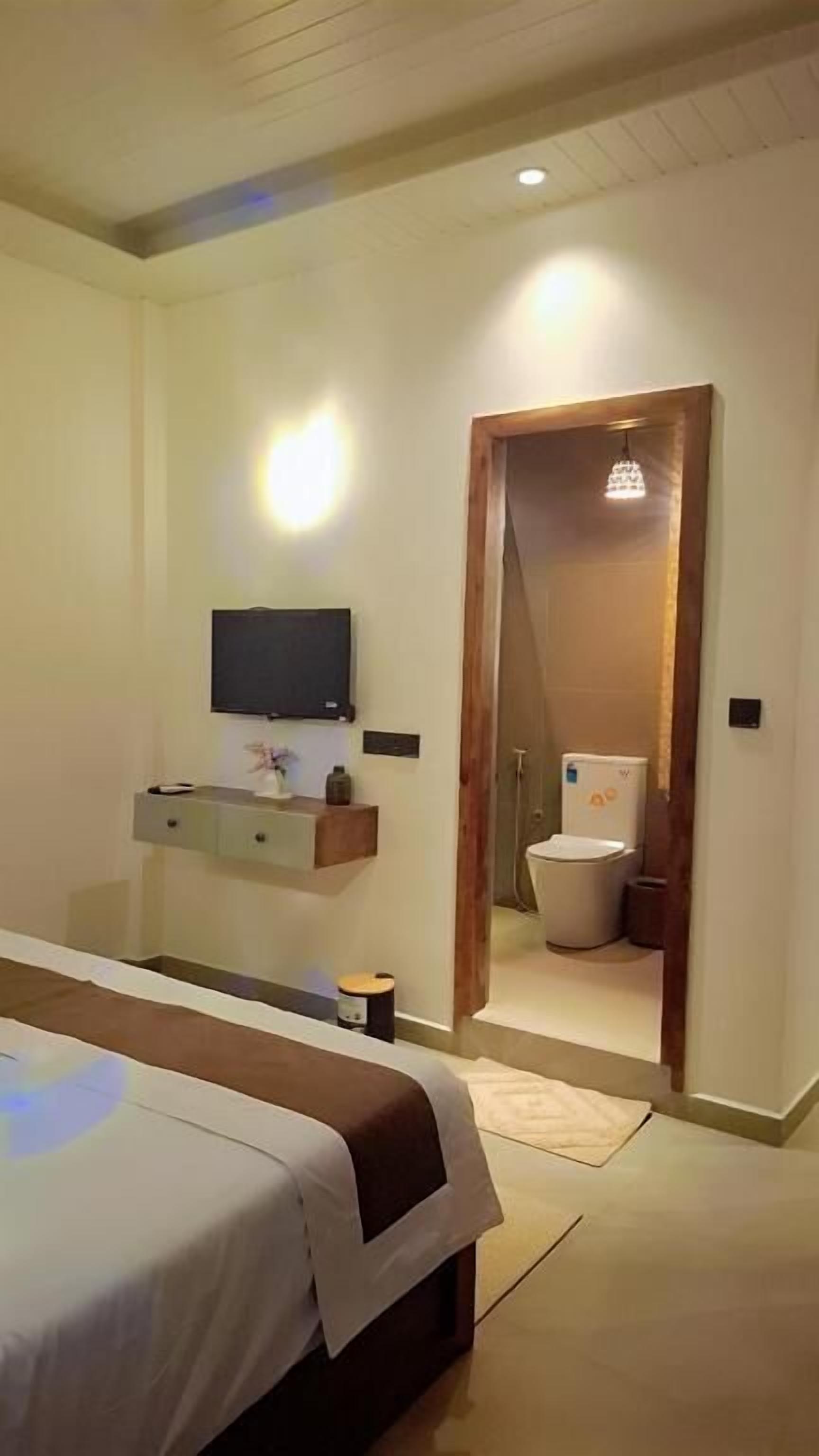 Ein Schlafzimmer mit einem Bett, einem an der Wand montierten Fernseher, einer Holztür zu einem Badezimmer und einem kleinen Tisch mit einer Vase.
