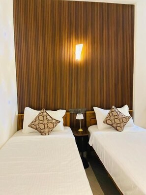 A hotel room with two beds, a wooden headboard, a nightstand with a lamp, and a wall-mounted light. - Secret Gems Resort (Arugambay)