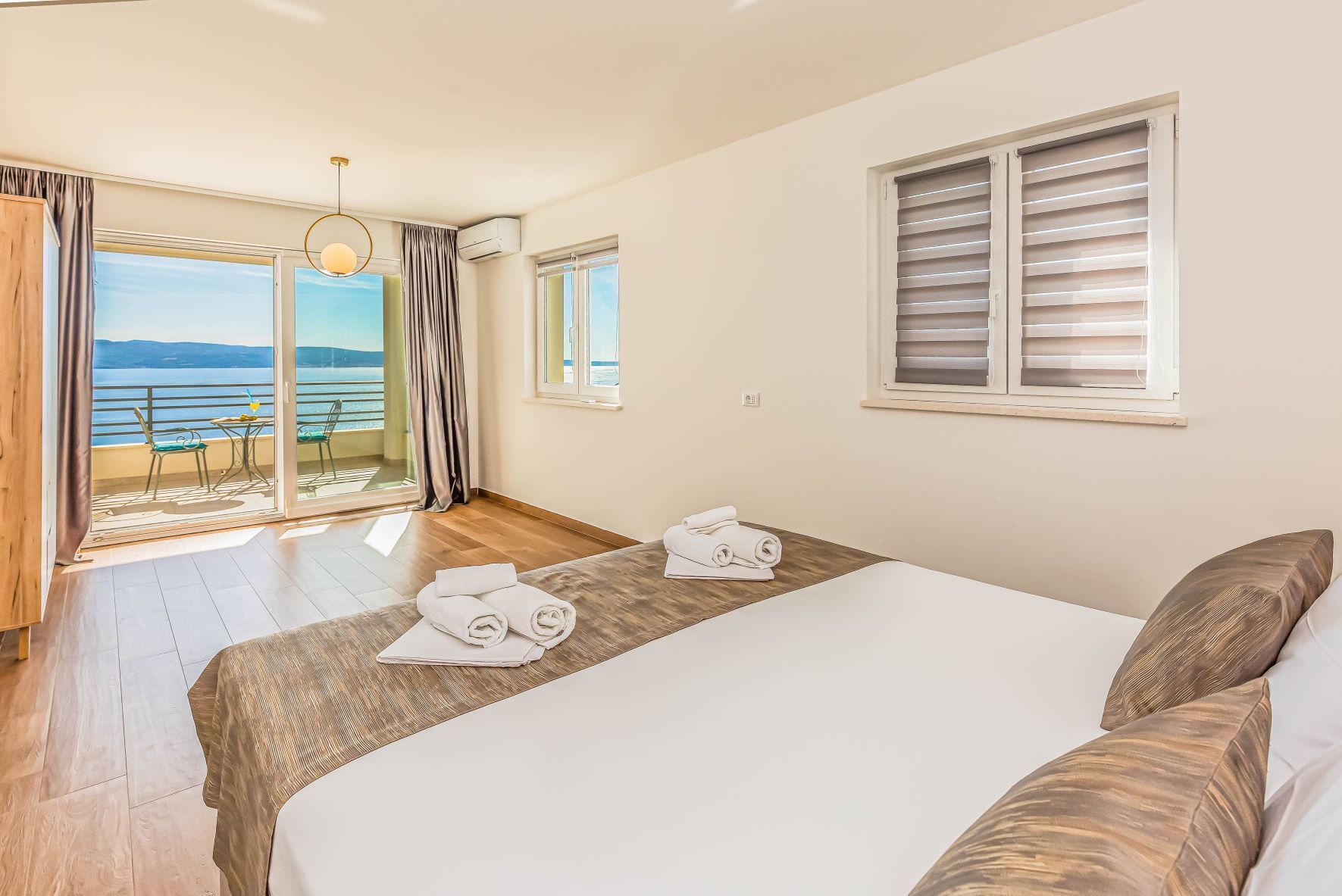 A hotel room with a bed, a table with towels, a balcony with a view, and a window with blinds.