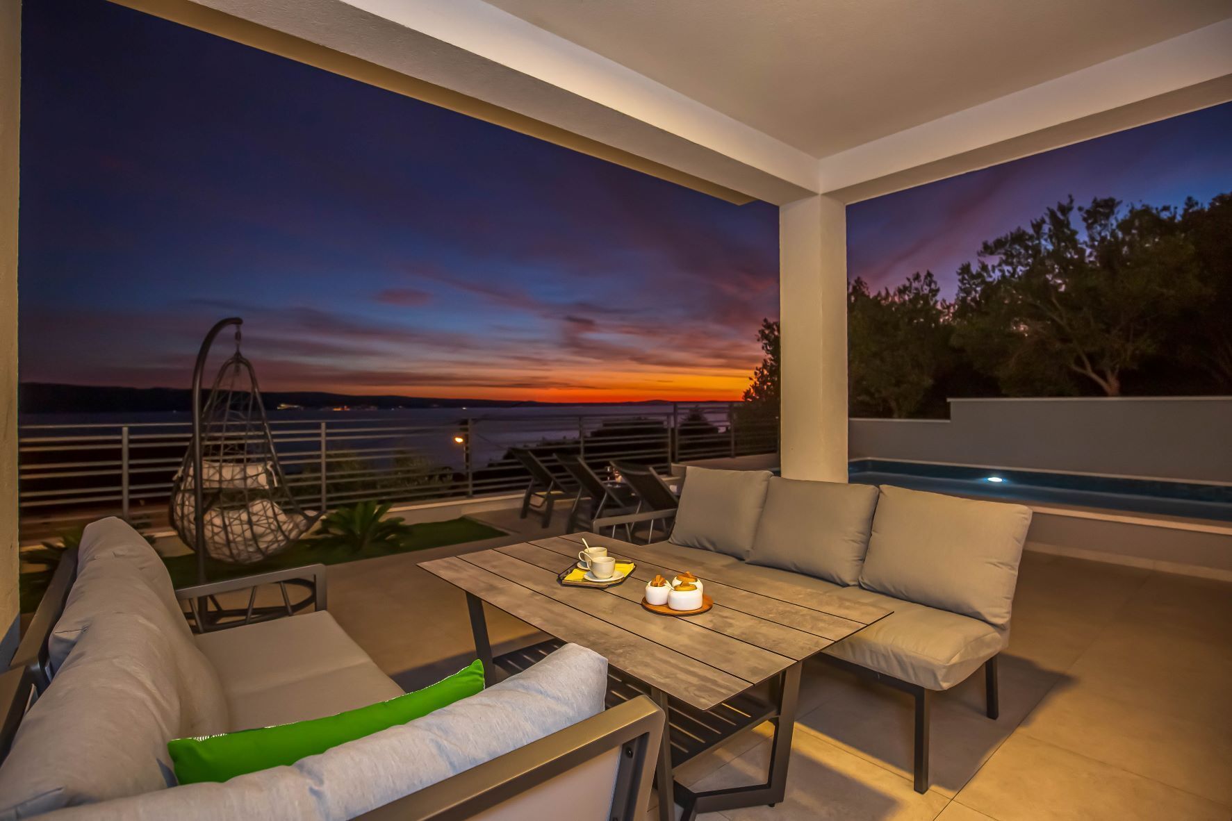 A modern outdoor seating area with a view of the sunset over the water, a pool, and a dining table with a candle and a cup.