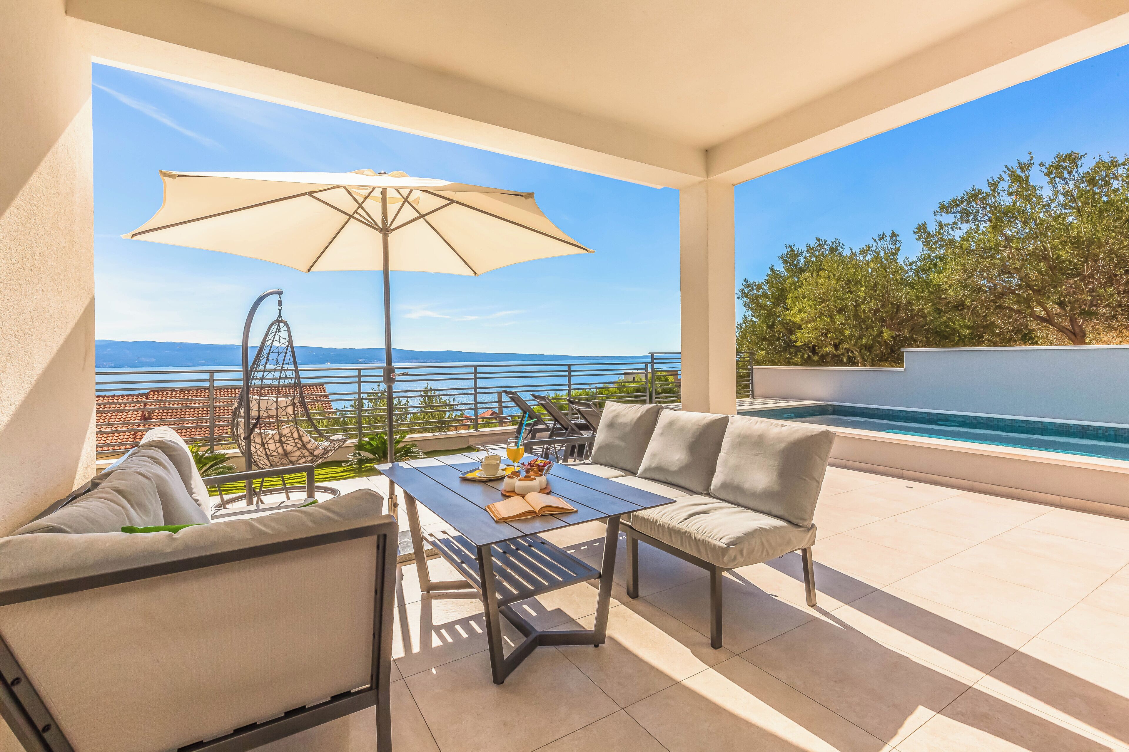 A terrace with a view of the sea, a swing chair, a table with a book and a drink, and a sofa.