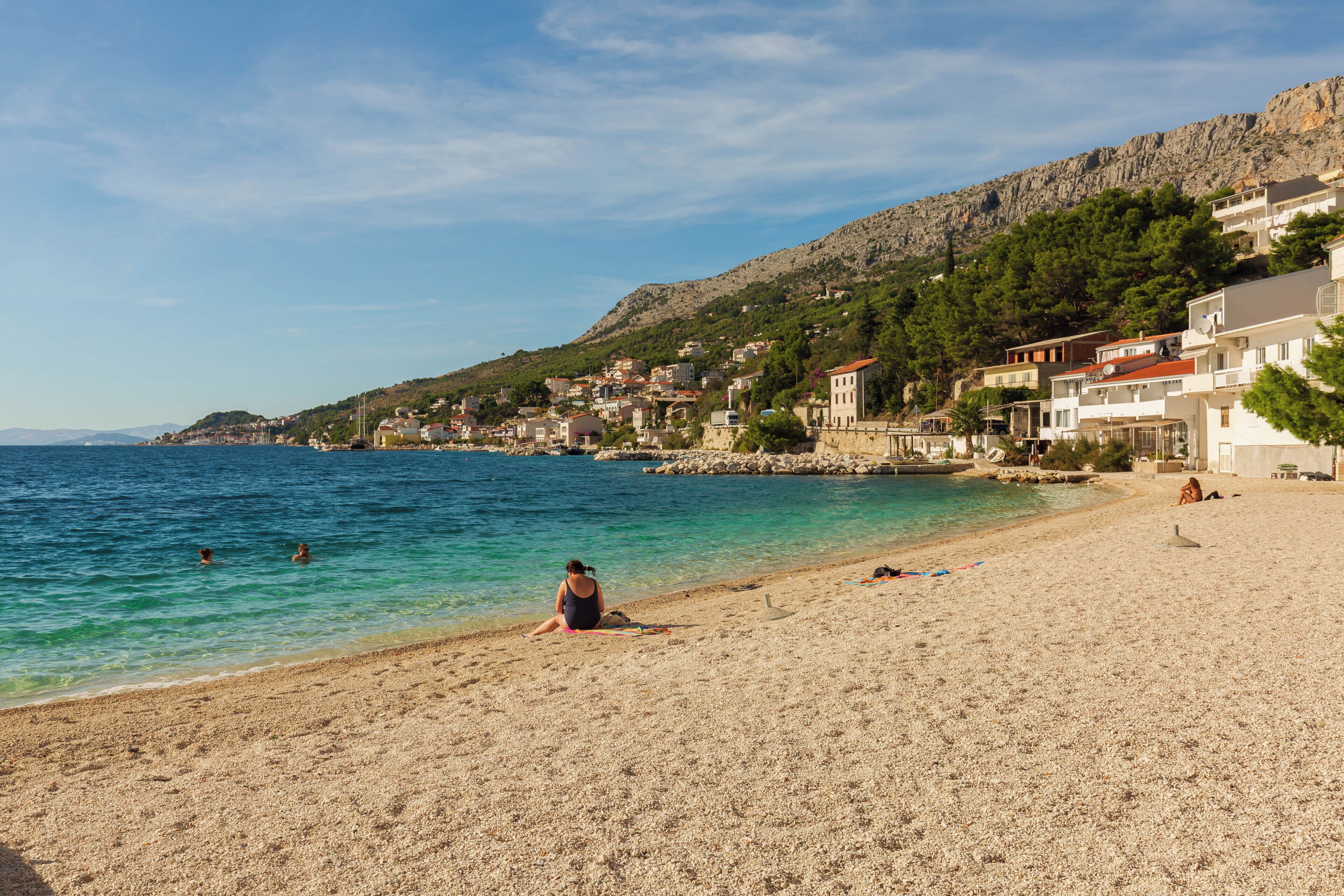A pebble beach with clear water, a person sitting on the sand, and buildings in the background.