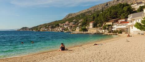 A pebble beach with clear water, a person sitting on the sand, and buildings in the background.