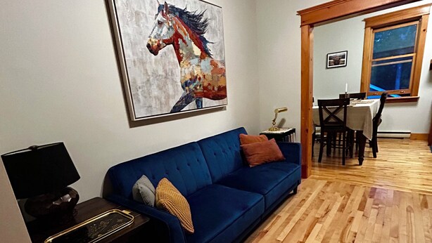 Una habitación con un sofá azul, suelo de madera, un cuadro de un caballo, una mesa con sillas y una ventana.
