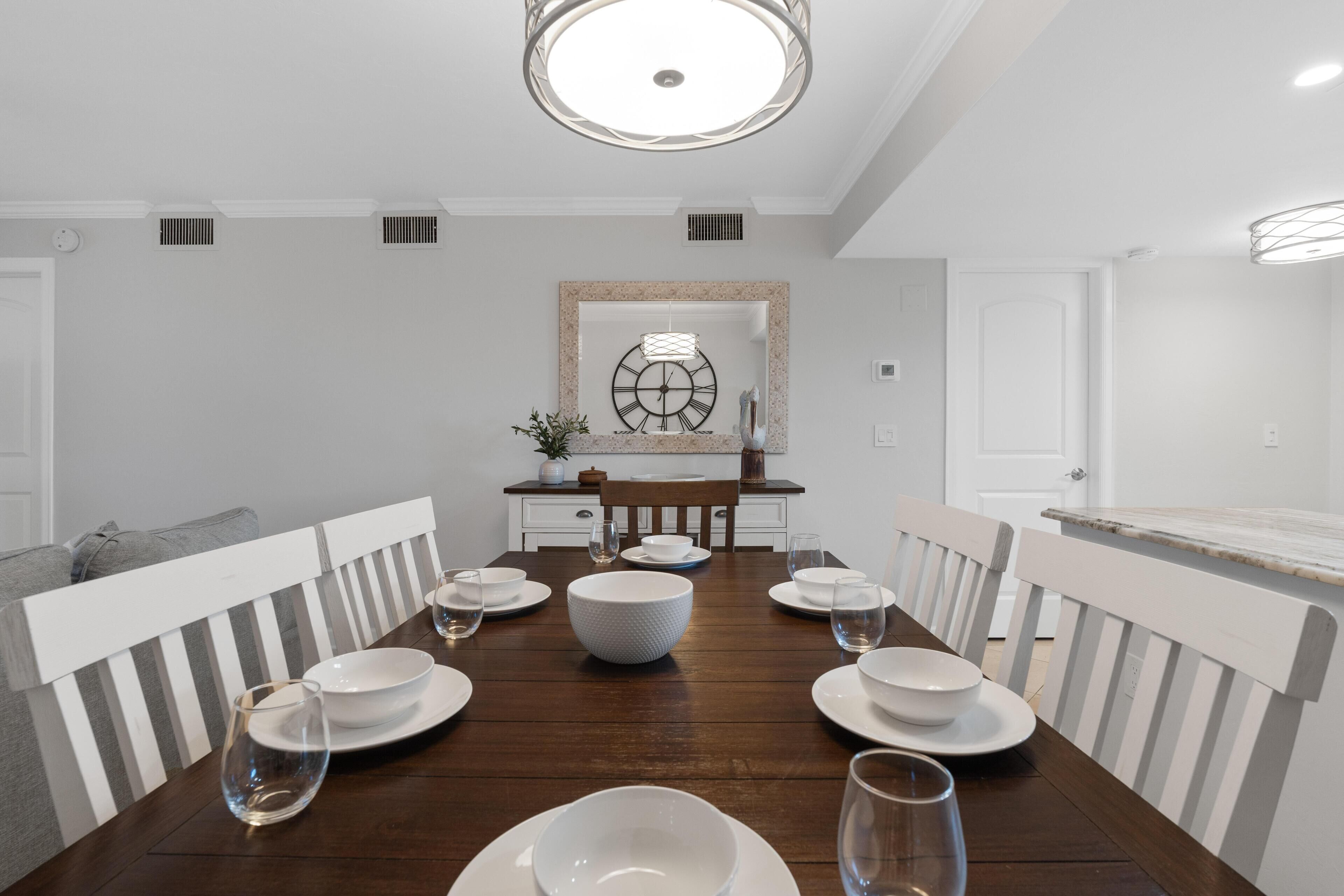 A dining room with a wooden table set for four, white chairs, and a large wall clock.