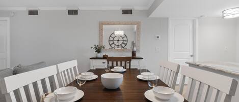A dining room with a wooden table set for four, white chairs, and a large wall clock.