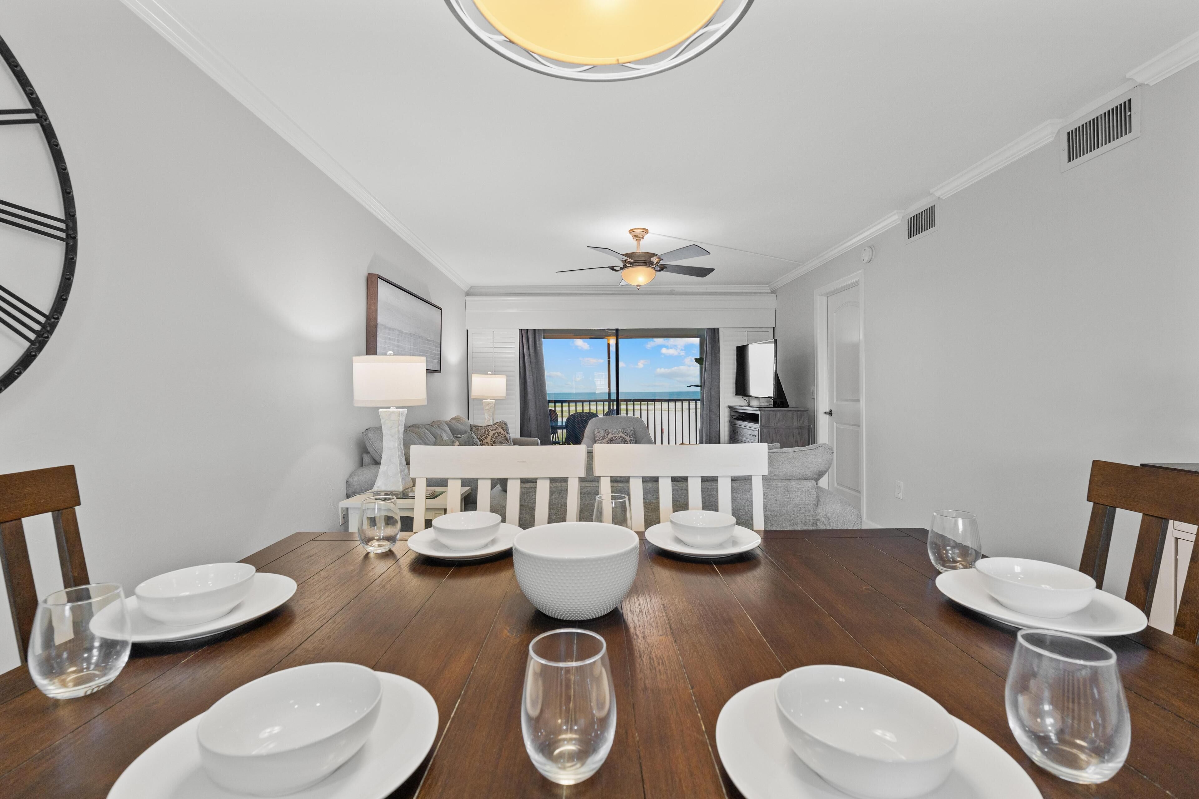 Comedor con mesa de madera dispuesta para seis personas, ventilador de techo y vista al océano a través de un ventanal.