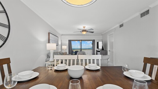 A dining area with a wooden table set for six, a ceiling fan, and a view of the ocean through a large window. - Carlos Pointe #213 Paradise Pointe (Fort Myers Beach)
