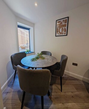 A dining area with a round table, four chairs, a window with a view, and a framed artwork on the wall. - City Centre Two Bedroom Apartment (Liverpool)