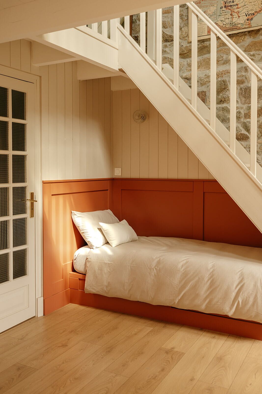 Une chambre avec un lit simple, un plancher en bois, une porte blanche avec une fenêtre et un escalier avec un mur en pierre.