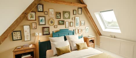 A bedroom with a sloped ceiling, a bed with multiple pillows, a nightstand with a lamp, and a wall adorned with framed pictures.