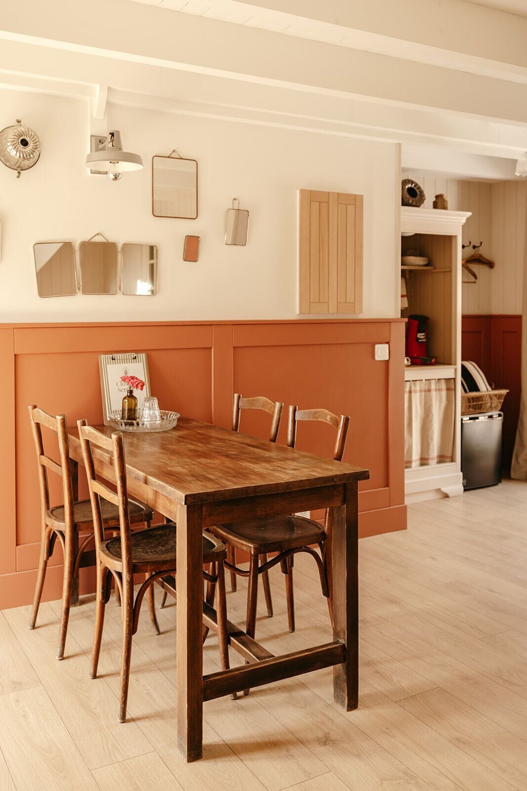A dining area with a wooden table and chairs, a wall-mounted light fixture, and a cabinet with a mirror and decorative items.