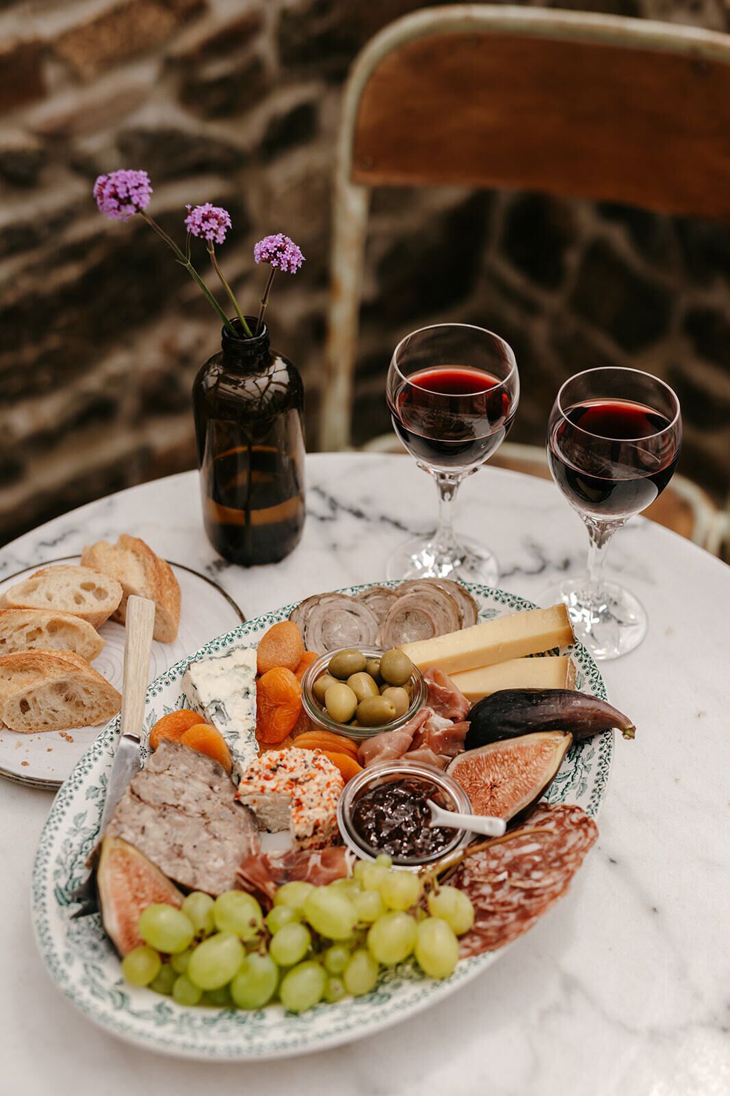 A charcuterie board with various meats, cheeses, and fruits, accompanied by two glasses of red wine, on a marble table.
