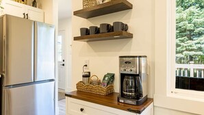 Una cocina con un refrigerador de acero inoxidable, repisas de madera con tazas, una cafetera y una cesta con objetos sobre la encimera.
