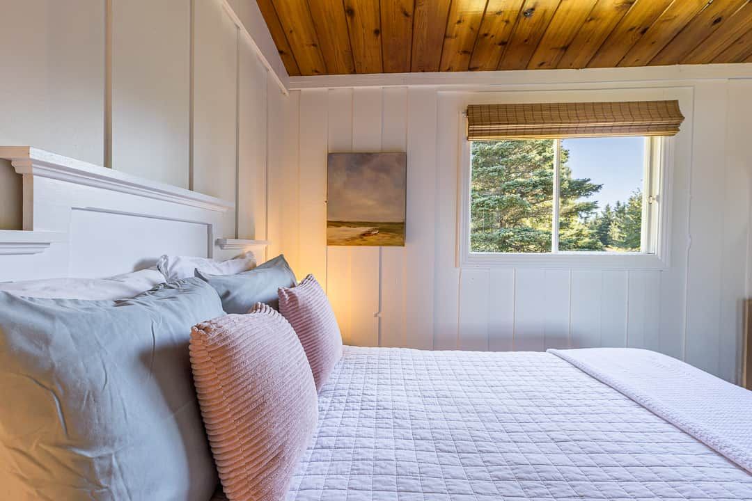 Quarto com cama, almofadas, um quadro e janela com vista para árvores.