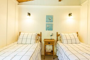 A room with two beds, each with a wooden headboard, a nightstand between them, and a wall-mounted light fixture. - Seawood Cottage on the Shore (North Rustico)