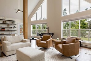 A spacious living room with a high ceiling, large windows, a piano, and a modern fireplace. - Lupin Lane Beach Retreat (Stratford)