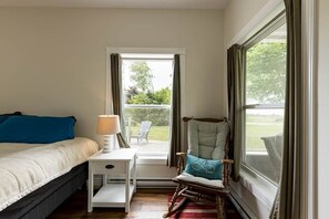 Et soveværelse med en seng, et natbord, en lampe, en gyngestol og et vindue med gardiner.