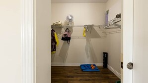 Een smalle gang met een houten vloer, een witte deur, een ventilator aan de muur en een plank met opgevouwen kleding.