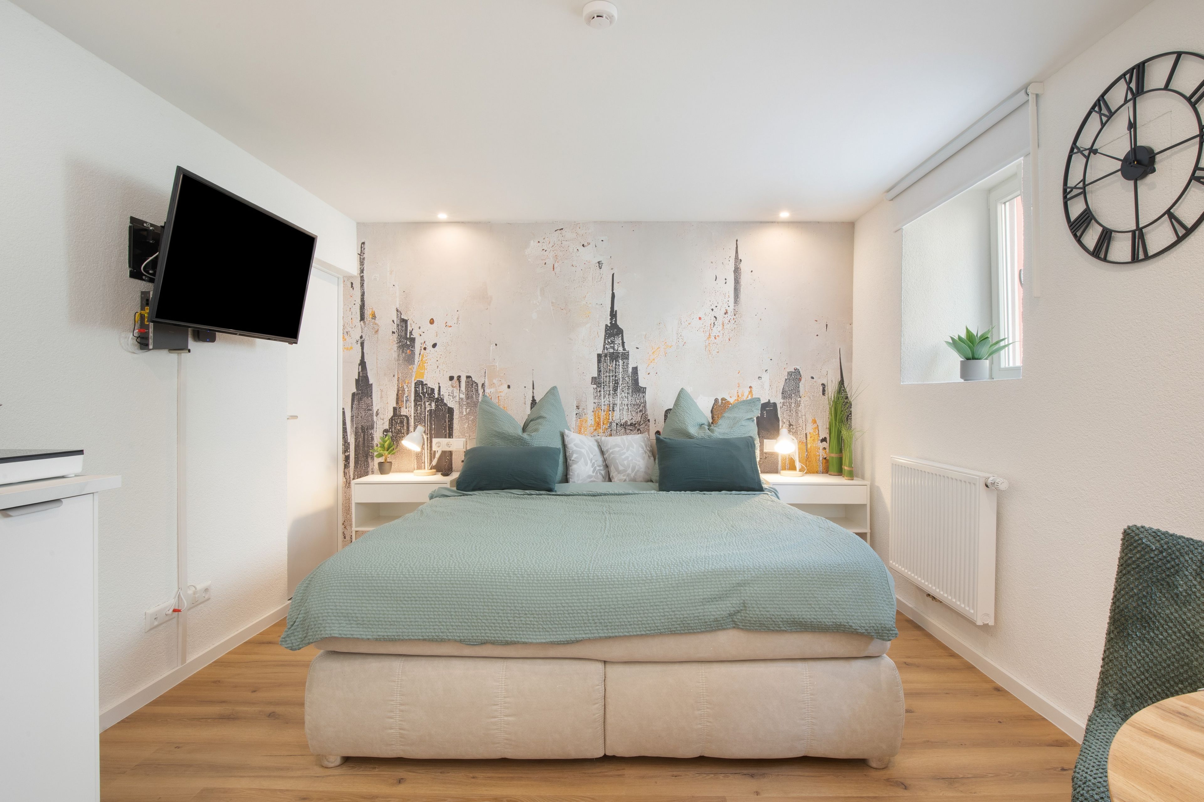 A bedroom with a bed, a wall-mounted TV, a cityscape mural, a clock, and a window with a plant.