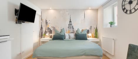 A bedroom with a bed, a wall-mounted TV, a cityscape mural, a clock, and a window with a plant.