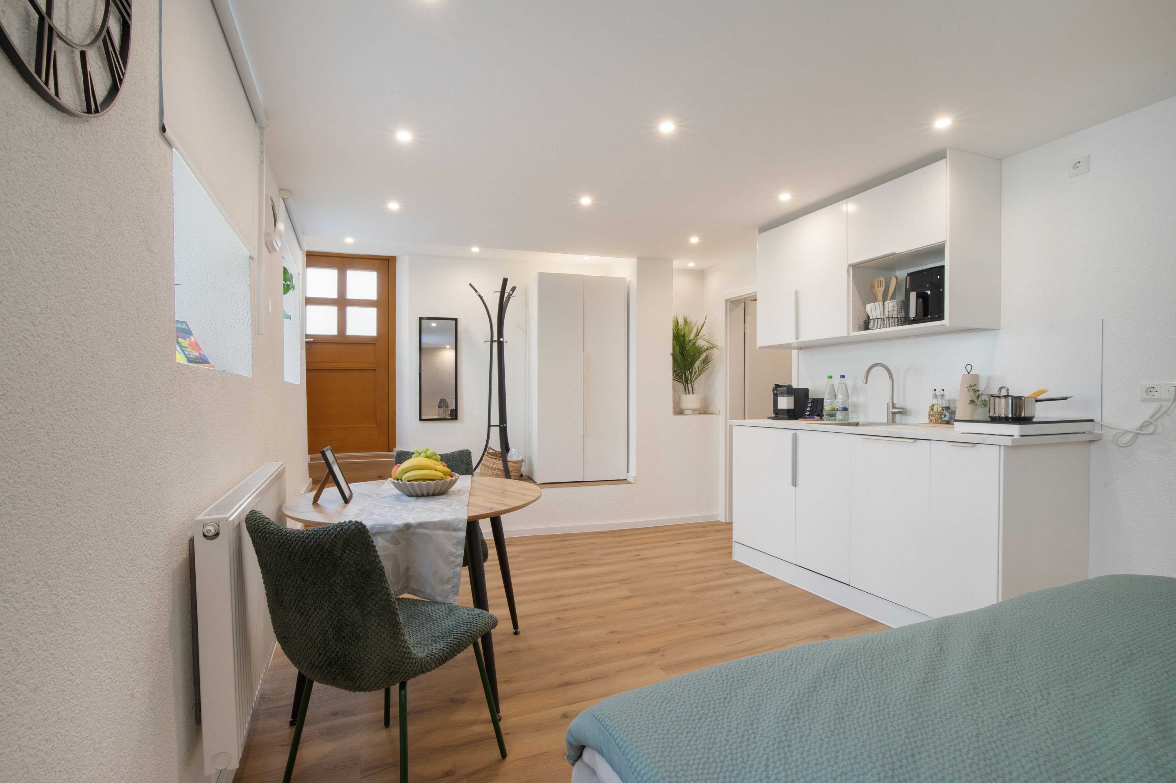 Een compacte keuken met witte kastjes, een eethoek met een tafel en stoelen, en een bed op de voorgrond.