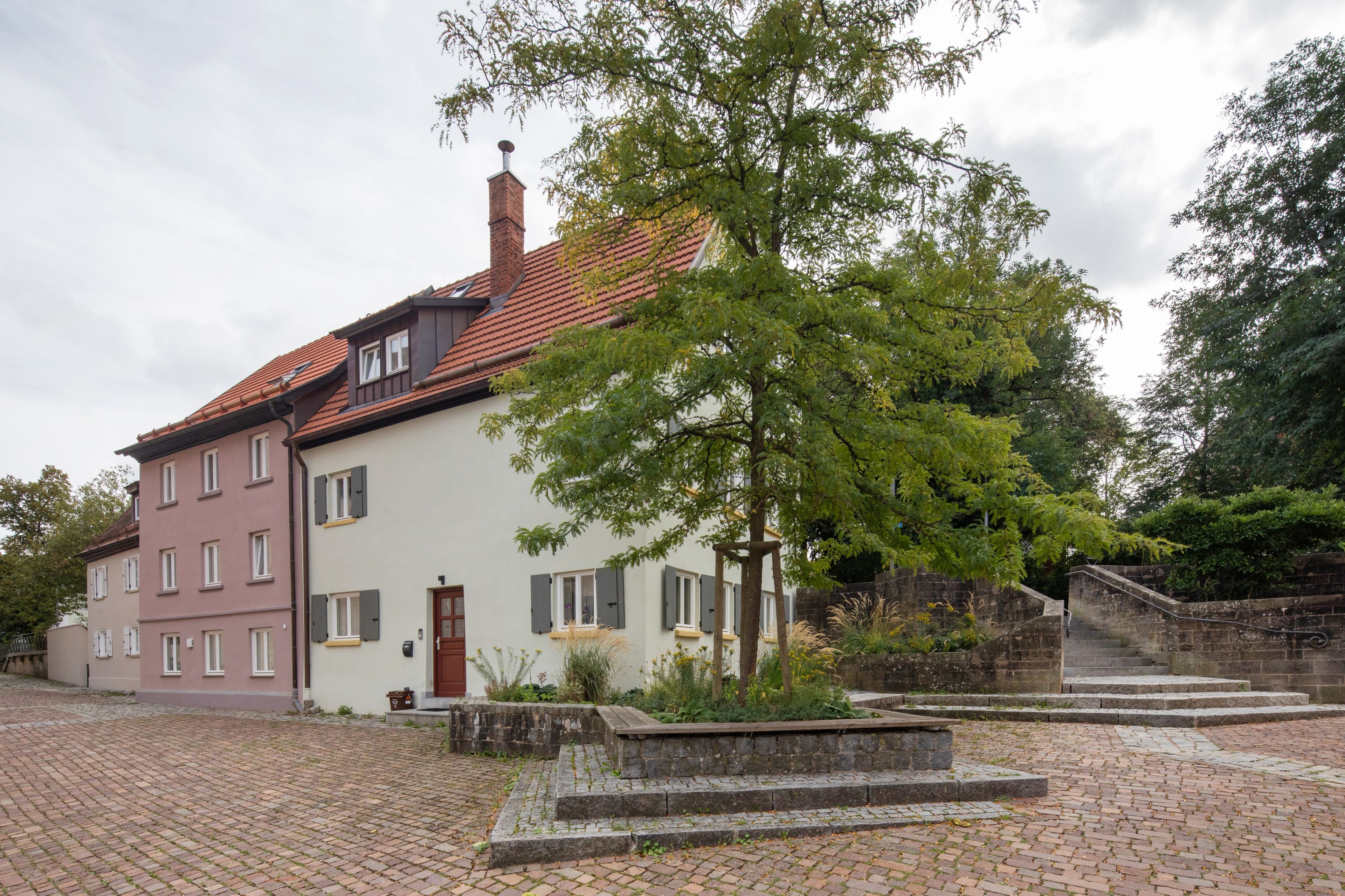 A two-story building with a red-tiled roof, a small garden with a tree, and a stone staircase leading to a door.