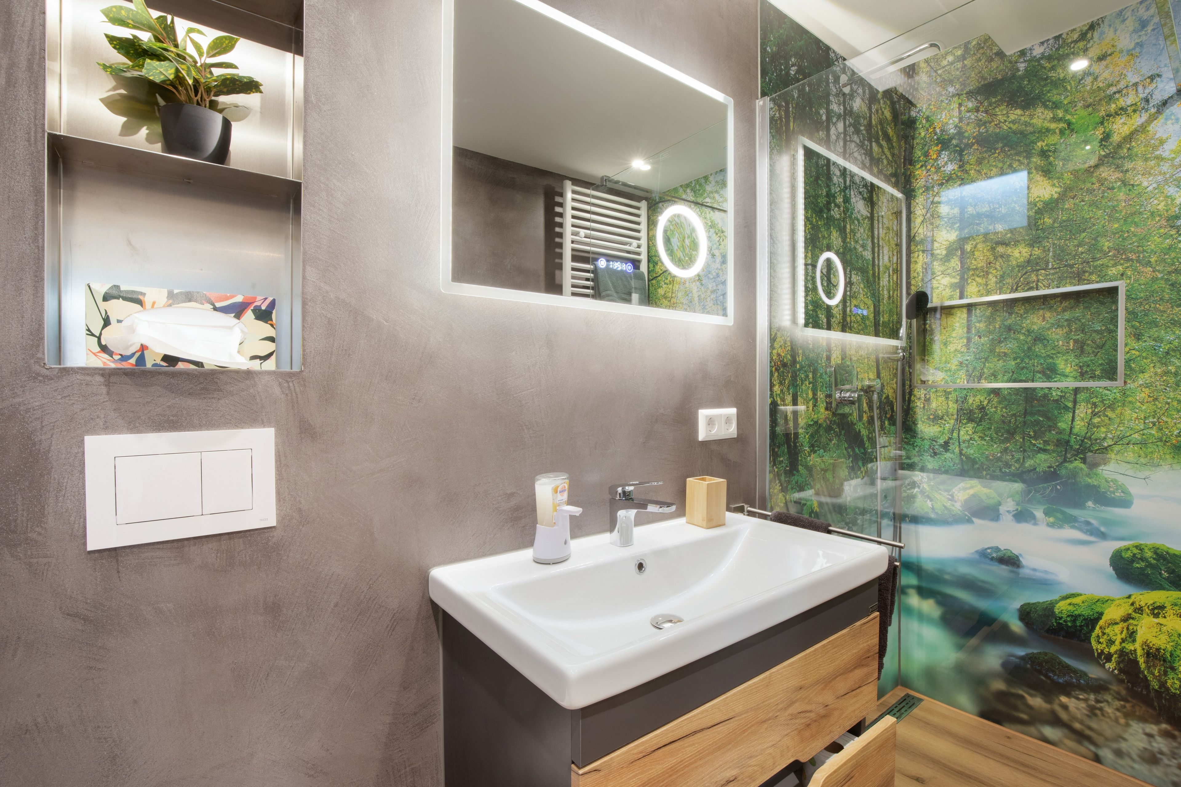 Ein Badezimmer mit einem weißen Waschbecken, einem Spiegel und einer Duschkabine aus Glas mit Blick auf einen Wald.