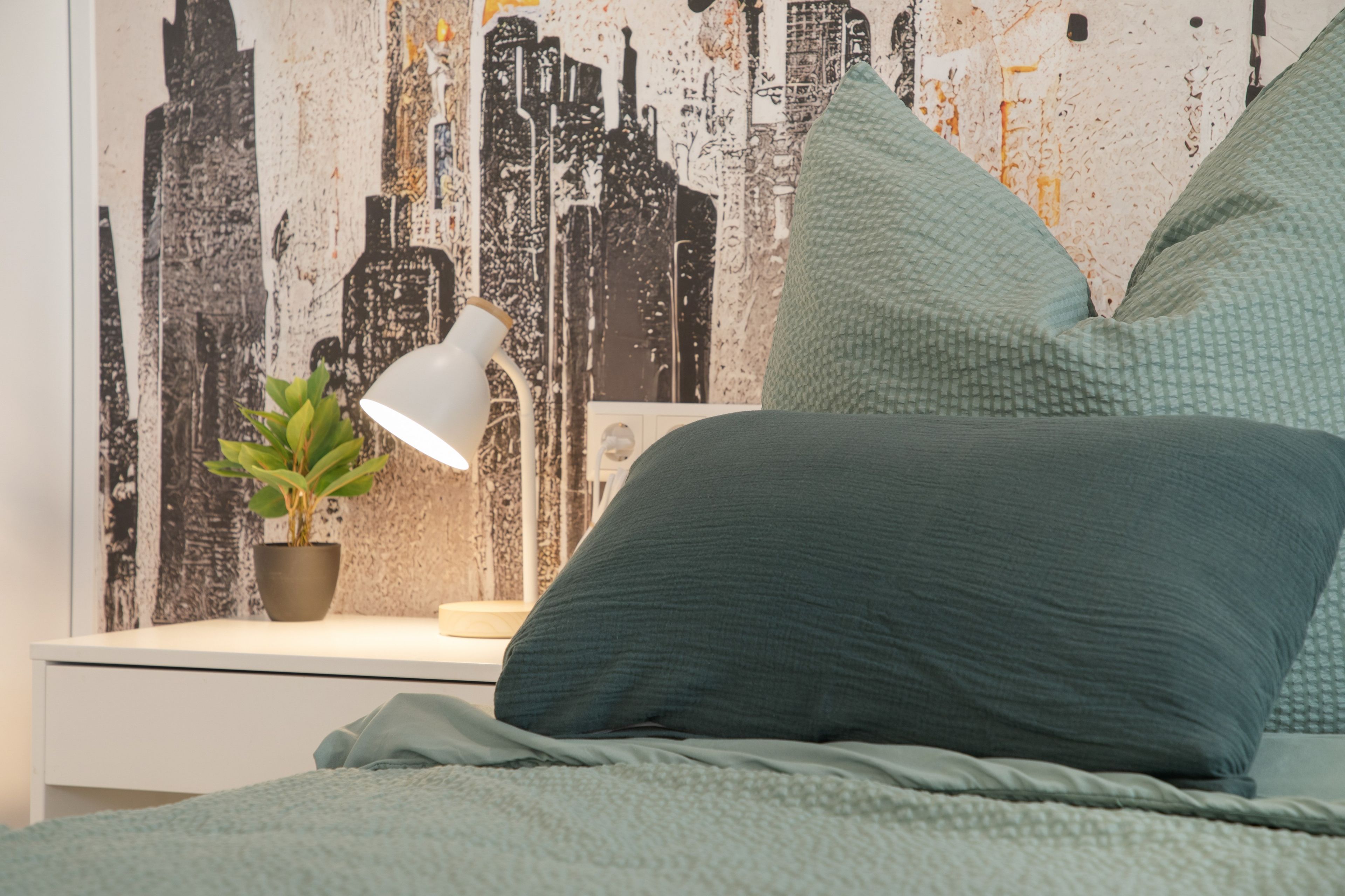 Ein Bett mit grüner Decke und Kissen, ein Nachttisch mit Lampe und Topfpflanze sowie eine Wand mit Stadtbild-Mural.