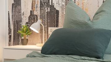 Ein Bett mit grüner Decke und Kissen, ein Nachttisch mit Lampe und Topfpflanze sowie eine Wand mit Stadtbild-Mural.