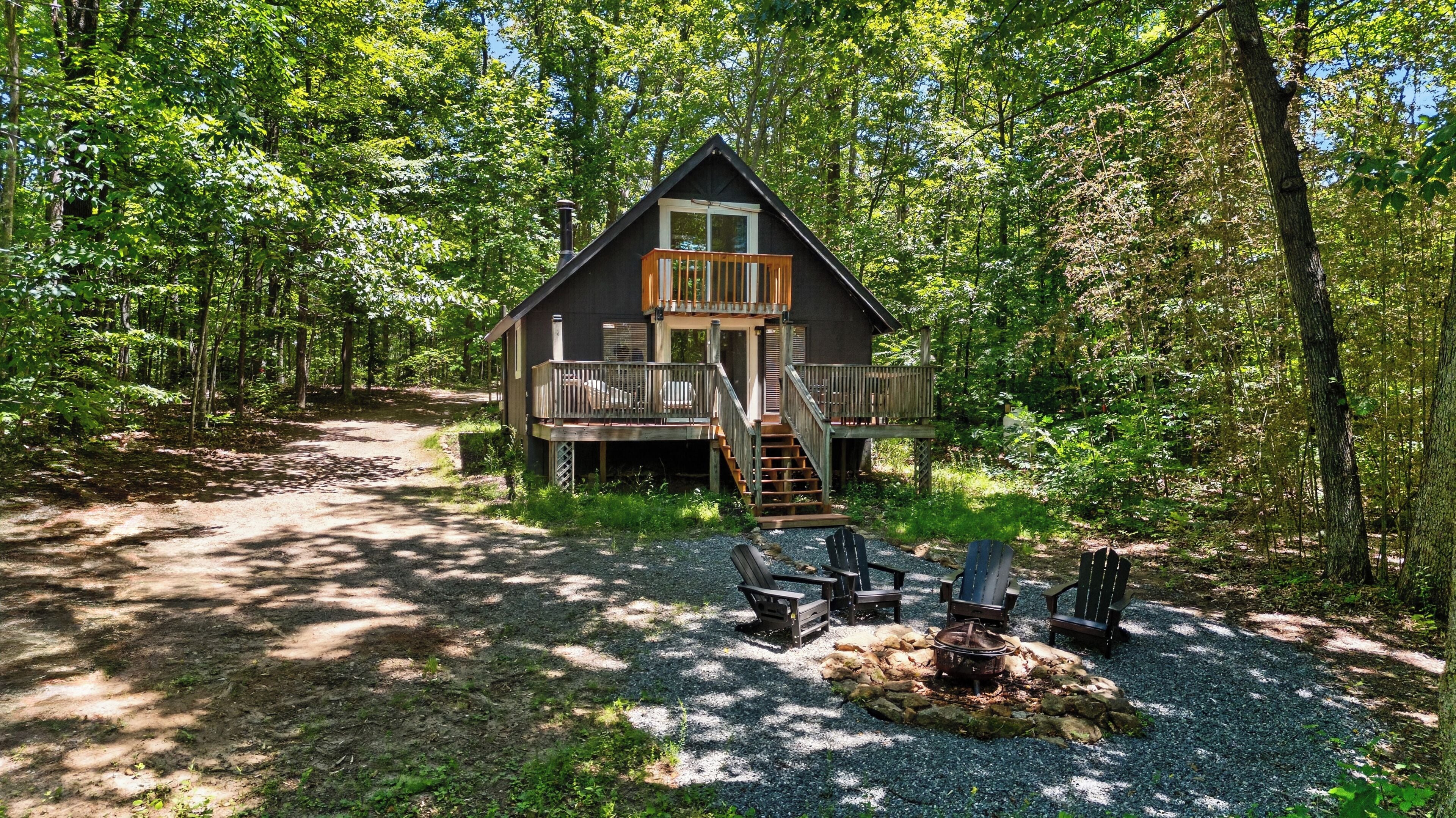 Une cabane dotée d’une terrasse, entourée d’arbres, et un foyer avec des chaises.