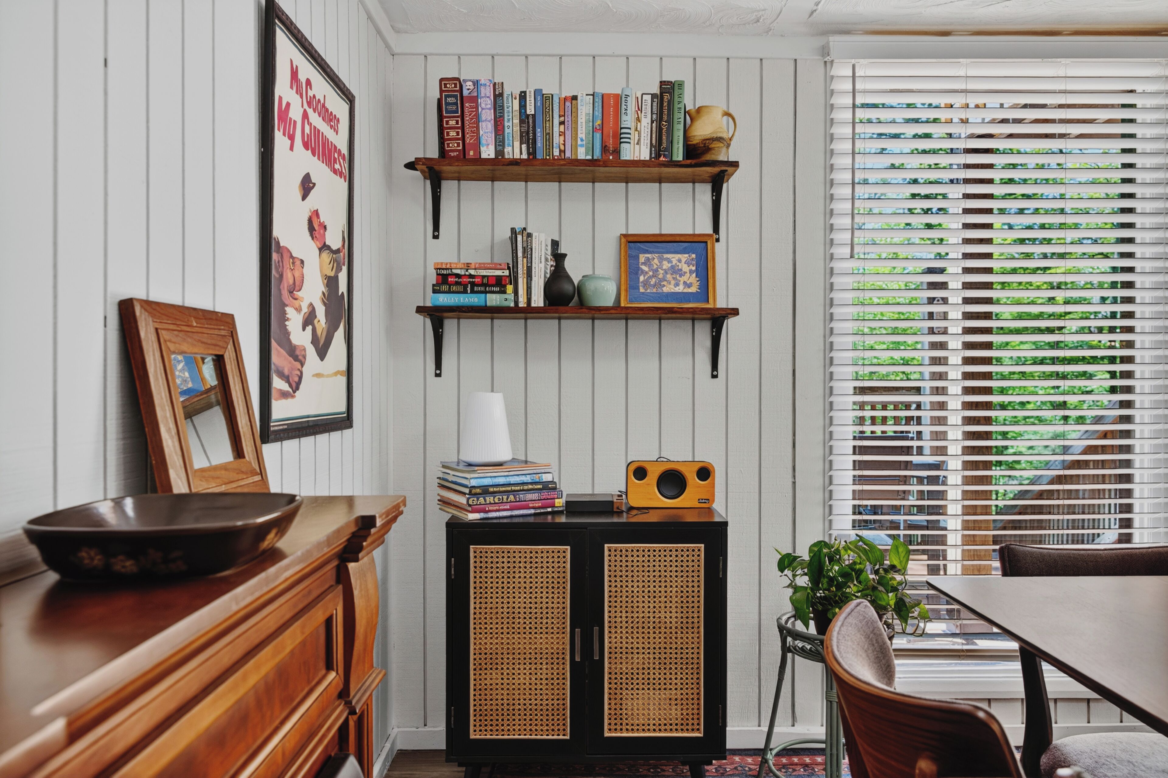 Une pièce comprenant une table à manger en bois, une bibliothèque remplie de livres et d’objets décoratifs, un tableau encadré et une fenêtre avec des stores.