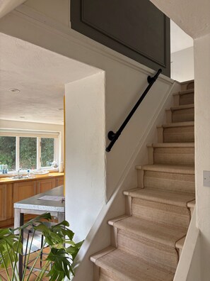A staircase with a handrail leading to a landing, a kitchen area with wooden cabinets, and a window with a view of greenery. - Cosy & private cottage nestled in the Wicklow Mountains. Pet friendly! (Tinahely)