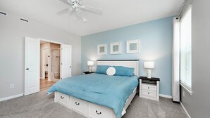 Un dormitorio con cama, mesitas de noche, ventilador de techo y una puerta que da a otra habitación.