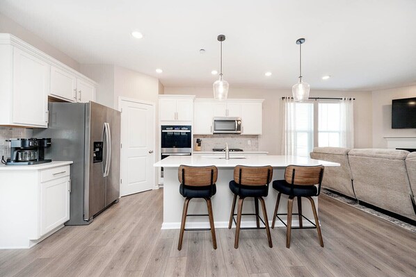 A modern kitchen with white cabinets, stainless steel appliances, a central island with bar stools, and a flat-screen TV mounted on the wall.