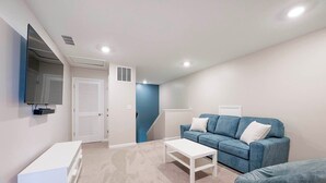 Una habitación con sofá azul, una mesita de centro blanca, un televisor montado en la pared y una puerta que da a otra estancia.