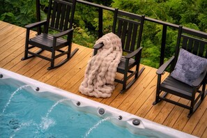 A wooden deck with a hot tub and two rocking chairs, one with a blanket. - SwimSpa/HotTub/GameRoom/FirePit&Yard/Peloton/LUXE (Gatlinburg)