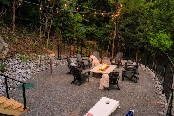 An outdoor seating area with a fire table surrounded by chairs, a wooden deck, and a gravel ground. - SwimSpa/HotTub/GameRoom/FirePit&Yard/Peloton/LUXE (Gatlinburg)