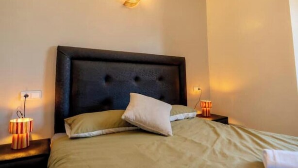 A bedroom with a tufted headboard, a bed with beige bedding, two bedside lamps, and a wall-mounted light fixture. - Bright Apartment & Rooftop Terrace (Essaouira)