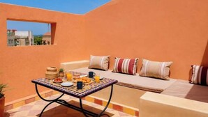 A sunlit outdoor seating area with a table set for breakfast, a cushioned sofa with striped cushions, and a view of buildings in the background. - Bright Apartment & Rooftop Terrace (Essaouira)