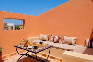 Uma área externa ensolarada com espaço para sentar, uma mesa posta para café da manhã, um sofá acolchoado com almofadas listradas e vista para edifícios ao fundo.